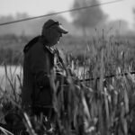 Black and white photo of an adult man fishing amidst tall reeds by a lake in a serene setting.